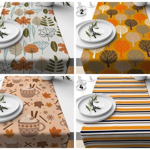May include: Four different table runner designs. The first runner features a white background with orange, brown, and green leaves. The second runner features a yellow background with orange, grey, and black trees. The third runner features a white background with orange, brown, and white leaves and a brown and white mug design. The fourth runner features a white and yellow striped design with orange and black stripes.