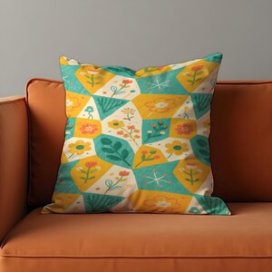Puede incluir: Cojín cuadrado con un estampado floral geométrico. El diseño presenta formas amarillas, turquesas y blanquecinas llenas de flores y hojas estilizadas. El cojín está sobre un sofá naranja.