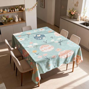 May include: A rectangular tablecloth with a light teal background, decorated with floral and egg designs in pink, white, and blue. The tablecloth is on a wooden table, surrounded by beige chairs, creating a spring or Easter theme.