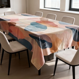 May include: A rectangular tablecloth with an abstract design. The pattern features large, overlapping shapes in shades of peach, coral, blue, and navy, with dotted and striped details. The tablecloth is draped over a dining table, surrounded by chairs.