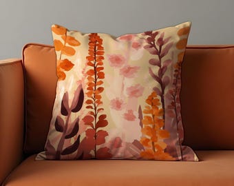 Op de afbeelding: Vierkant sierkussen met een bloemenontwerp. Het kussen heeft een crèmekleurige achtergrond met aquarel illustraties van oranje, rode en roze bloemen en bladeren. Het kussen ligt op een oranje bank.