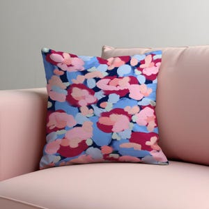 May include: Square decorative pillow with a floral pattern. The pillow features a blue background with pink, red, and light pink brushstrokes. The pillow is placed on a pink sofa. The pillow is a home decor item.