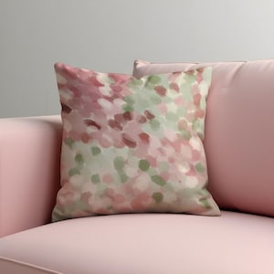 Puede incluir: Un cojín decorativo cuadrado con un diseño de estilo acuarela. El patrón presenta una mezcla de pinceladas rosas, verdes y burdeos. El cojín está colocado sobre un sofá rosa, creando una estética suave y acogedora.
