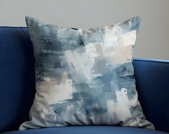 Può includere: Cuscino quadrato decorativo con un design astratto nei toni del blu, grigio e bianco. Il cuscino è posto su un divano blu scuro, creando un contrasto di colori. Il design del cuscino assomiglia a pennellate.