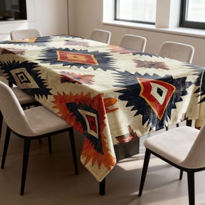 May include: A rectangular tablecloth with a cream-colored background and a geometric pattern in shades of navy blue, orange, and red. The pattern features diamond and star-like shapes. The tablecloth is draped over a rectangular table.
