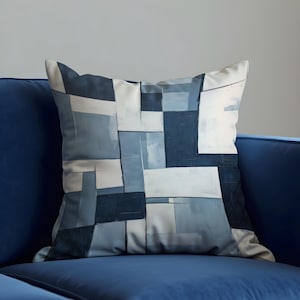 Può includere: Cuscino quadrato con un design geometrico astratto nei toni del blu, grigio e bianco. Il cuscino è posto su un divano blu, creando una palette di colori coerente.
