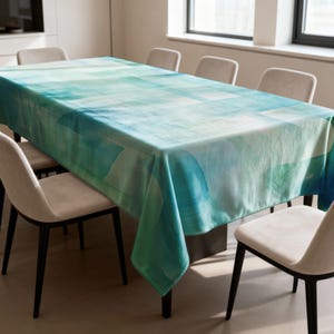 Puede incluir: Un mantel rectangular con un diseño abstracto de acuarela en tonos azules y verdes. El mantel está sobre una mesa oscura, rodeada de sillas beige. El diseño presenta formas suaves y difuminadas.