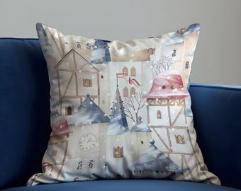 Könnte beinhalten: Quadratisches Zierkissen mit einem Aquarelldruck einer verschneiten Dorflandschaft. Das Design zeigt Gebäude mit roten, blauen und beigen Dächern und einen Uhrturm. Das Kissen liegt auf einem blauen Sofa.