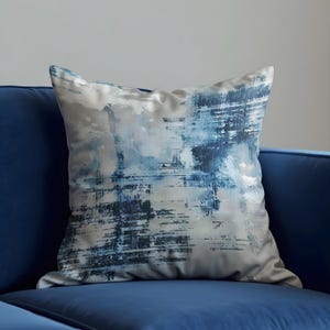 Könnte beinhalten: Ein quadratisches Zierkissen mit einem abstrakten Design in Blau- und Weißtönen. Das Kissen liegt auf einem blauen Sofa. Das Design weist pinselstrichartige Muster auf, die ein strukturiertes Aussehen erzeugen. Der Stoff des Kissens hat einen weichen, samtigen Glanz.