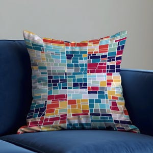 Puede incluir: Un cojín decorativo con un colorido diseño geométrico. El diseño presenta un mosaico de formas rectangulares en tonos de azul, rojo, naranja, amarillo y gris, con contornos blancos. El cojín está sobre un sofá azul.