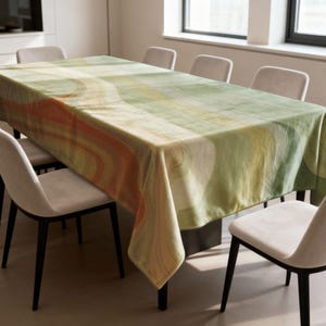 May include: A rectangular tablecloth with an abstract design in shades of green, orange, and yellow covers a dining table. The pattern features curved lines and shapes. The table is surrounded by six chairs with white seats and black legs.