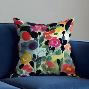 May include: Square throw pillow with a watercolor floral design. The pillow features a vibrant mix of red, pink, yellow, and black flowers with green leaves against a light background. The pillow is placed on a blue sofa.