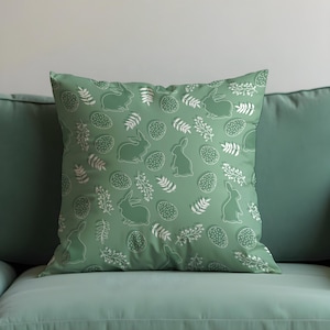 May include: A square, sage green cushion featuring a repeating pattern of white Easter eggs, rabbits, and sprigs. The cushion is placed on a matching green sofa, creating a decorative look.