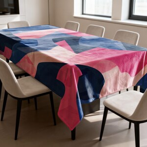 May include: A rectangular tablecloth with a modern abstract design. The pattern features overlapping shapes in shades of pink, blue, and navy. The tablecloth is draped over a dining table, with chairs visible around it. The overall theme is home decor.