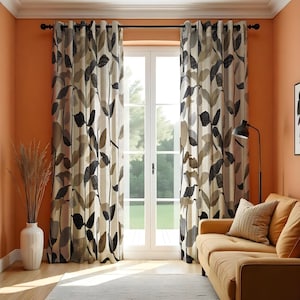 May include: Curtains with a leaf pattern in black, brown, and beige. The curtains are hanging in front of a window. A tan sofa and a floor lamp are visible in the room. The walls are painted in a warm orange colour. The curtains are made of fabric.