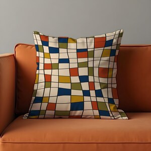 May include: Square throw pillow with a colorful geometric pattern. The design features a grid of squares in shades of beige, blue, orange, green, and yellow, outlined with black lines. The pillow is placed on an orange sofa.