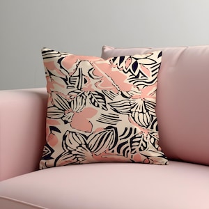 Op de afbeelding: Vierkant decoratief kussen met een bloemenpatroon. Het kussen heeft een crèmekleurige achtergrond met roze en marineblauwe bloemmotieven. Het kussen ligt op een roze bank.