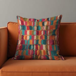 May include: Square decorative pillow with a colorful abstract pattern. The pillow features a repeating design of vertical, oval shapes in shades of pink, orange, teal, and white. The pillow is resting on an orange sofa.