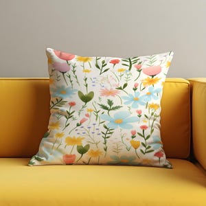 May include: Square throw pillow with a floral pattern. The pillow features a white background with a repeating design of colorful flowers in shades of pink, yellow, and blue. Green leaves and stems complete the design. The pillow is on a yellow couch.