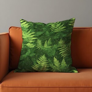 May include: Square throw pillow featuring a vibrant green fern leaf pattern. The pillow's design showcases a dense arrangement of fern fronds in varying shades of green, creating a lush, natural aesthetic. The pillow is placed on an orange sofa.