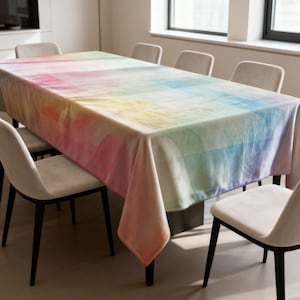 Puede incluir: Un mantel rectangular con un diseño de arcoíris en acuarela. Los colores se mezclan del rosa al amarillo, al azul y al verde. El mantel está colocado sobre una mesa rectangular, rodeada de sillas.