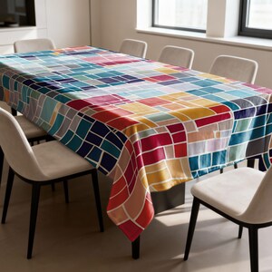 Op de afbeelding: Een rechthoekig tafelkleed met een kleurrijk geometrisch patroon. Het ontwerp heeft een raster van vierkanten in tinten rood, oranje, geel, blauw en grijs. Het tafelkleed ligt op een tafel met stoelen eromheen.