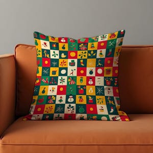 May include: Square throw pillow with a colorful Christmas-themed pattern. The design features a grid of squares in red, green, yellow, and white, each containing festive illustrations like snowmen, gifts, and snowflakes. The pillow is placed on an orange sofa.