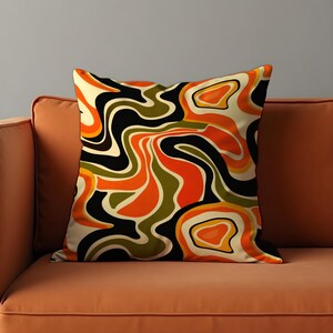 May include: A square cushion with a retro abstract design. The pattern features swirling shapes in orange, black, olive green, and cream colours. The cushion is placed on an orange sofa, creating a cohesive colour palette.