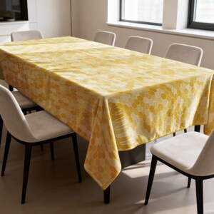 Peut inclure: Une nappe rectangulaire jaune avec un motif en nid d'abeille recouvre une table à manger. La nappe est drapée sur une table sombre, entourée de six chaises aux assises grises claires et aux pieds noirs. La pièce a un design moderne.