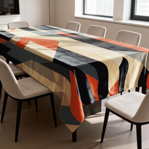 Pode incluir: Uma toalha de mesa retangular com um design geométrico. O padrão apresenta formas pretas, cinzentas, laranja e bege. A toalha de mesa é colocada sobre uma mesa de madeira, rodeada por cadeiras brancas.
