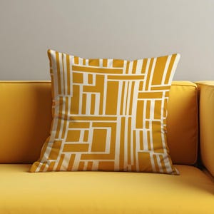 Può includere: Cuscino quadrato decorativo con un motivo geometrico giallo senape e crema. Il cuscino è posto su un divano giallo. Il motivo è composto da forme rettangolari e lineari, creando un design moderno. Aggiunge colore e stile.