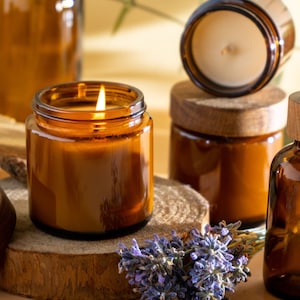 May include: A set of amber glass candle jars with wooden lids and a small amber glass bottle with a dropper. The candles are lit and the bottle is filled with a clear liquid. There are lavender flowers in the foreground.