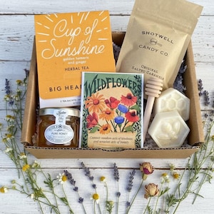 May include: A gift box filled with a variety of items, including a jar of honey, a packet of herbal tea, a bag of salted caramels, and two bee-shaped soaps. The box also contains a wooden honey dipper and a card with wildflower illustrations.
