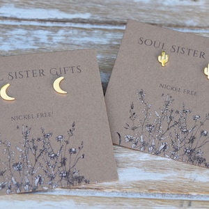 May include: Two pairs of gold earrings on brown cards. The first pair features a crescent moon design. The second pair features a cactus design. Both cards have the text "Soul Sister Gifts" and "Nickel Free".