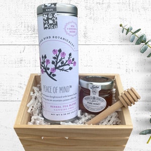 May include: A wooden gift box filled with white shredded paper, containing a tin of herbal tea blend labeled "Peace of Mind" and a small glass jar of honey with a wooden honey dipper. The tea tin features a floral design with pink flowers.