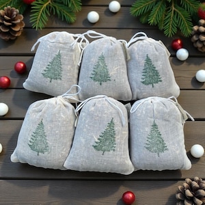 May include: Six gray linen drawstring bags with a green Christmas tree design are arranged on a wooden surface. The bags are filled and tied with white string. Decorative elements include pinecones, red and white ornaments, and evergreen branches.