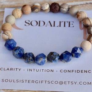 May include: A blue sodalite gemstone bracelet with brown and white jasper beads. The bracelet is on a white card with the text "Sodalite", "Clarity - Intuition - Confidence", and "Soulsistergiftsco@etsy.com".