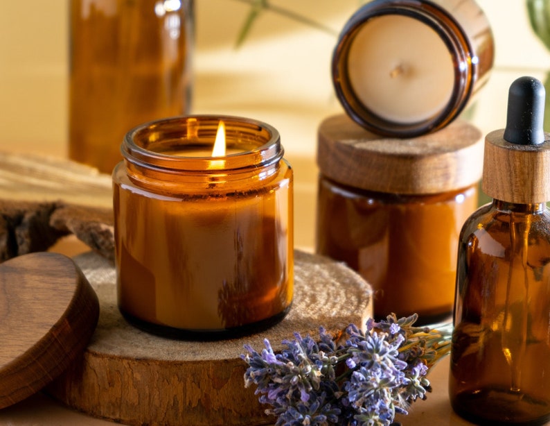 Puede incluir: Un conjunto de tarros de velas de vidrio &aacute;mbar con tapas de madera y una peque&ntilde;a botella de vidrio &aacute;mbar con un gotero. Las velas est&aacute;n encendidas y la botella est&aacute; llena de un l&iacute;quido transparente. Hay flores de lavanda en primer plano.