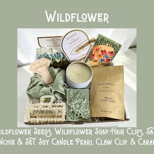 May include: A gift box with the word "WILDFLOWER" at the top. The box contains wildflower seeds, soap, hair clips, a satin scrunchie set, a soy candle, a pearl claw clip, and caramels. The box is filled with various items in shades of green and white.