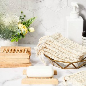 May include: A set of three wooden soap dishes stacked on top of each other. A bar of white soap is sitting on a wooden soap dish. A white mesh bag with a drawstring is sitting in a gold metal bowl. The mesh bag is made of natural fibers and is designed to hold soap.