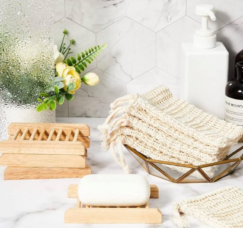 May include: A set of three wooden soap dishes stacked on top of each other. A bar of white soap is sitting on a wooden soap dish. A white mesh bag with a drawstring is sitting in a gold metal bowl. The mesh bag is made of natural fibers and is designed to hold soap.