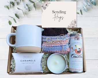 May include: A gift box filled with cozy items for self-care, including a white mug, brown and orange patterned socks, a white candle, a small box of caramels, and a tin of drinking chocolate. The box is decorated with shredded paper and a card that says "Sending Hugs".