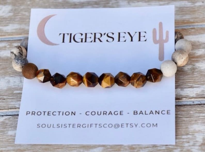 May include: A brown and gold tiger's eye gemstone bracelet with white beads. The bracelet is on a white card with the text "TIGER'S EYE" and "PROTECTION - COURAGE - BALANCE" and the website address "SOULSISTERGIFTSCO@ETSY.COM".