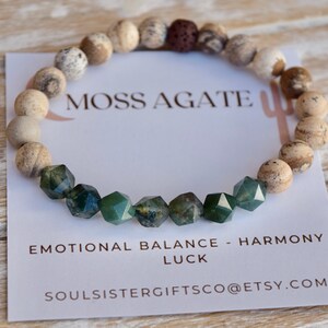 May include: A bracelet made with moss agate and brown and white jasper beads. The bracelet has a lava bead and the text "Moss Agate" is printed on a white card. The card also says "Emotional Balance - Harmony Luck" and "soulsistergiftsco@etsy.com".