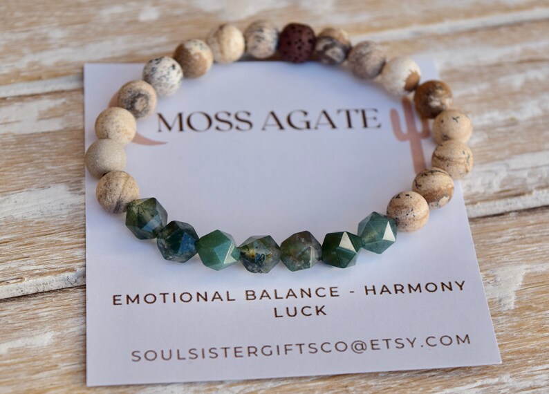 May include: A bracelet made with moss agate and brown and white jasper beads. The bracelet has a lava bead and the text "Moss Agate" is printed on a white card. The card also says "Emotional Balance - Harmony Luck" and "soulsistergiftsco@etsy.com".