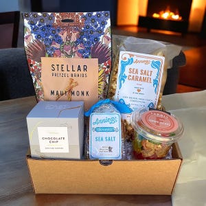 May include: A brown cardboard gift box filled with various snacks. The box contains a bag of pretzel braids, a bag of sea salt caramel popcorn, a bag of sea salt caramels, a container of chocolate chip cookies, and a container of gummy bears. The box is sitting on a table in front of a fireplace.