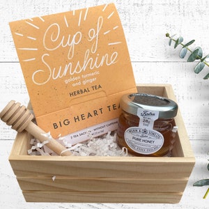 May include: A wooden crate filled with white shredded paper, a small jar of honey, a wooden honey dipper, and a package of herbal tea labeled "Cup of Sunshine" golden turmeric and ginger. The tea package also says "Big Heart Tea" and "2 tea sacks - without caffeine".