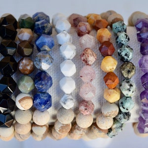 May include: A collection of 14 gemstone bracelets, each featuring a different type of stone. The bracelets are made with faceted beads and are arranged in a row on a white background. The stones include amethyst, rose quartz, tiger's eye, aventurine, and more.