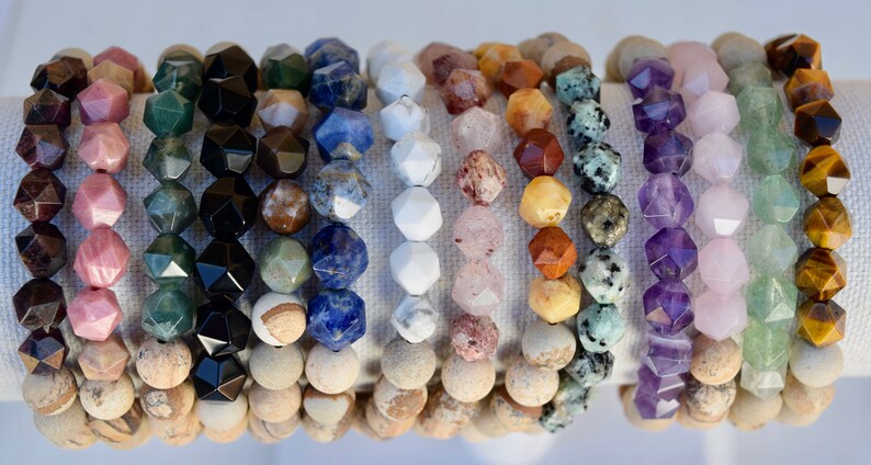 May include: A collection of 14 gemstone bracelets, each featuring a different type of stone. The bracelets are made with faceted beads and are arranged in a row on a white background. The stones include amethyst, rose quartz, tiger's eye, aventurine, and more.