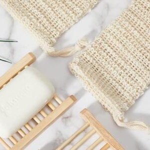 May include: Two white crocheted soap pouches with drawstrings and a white bar of soap in a wooden soap dish.